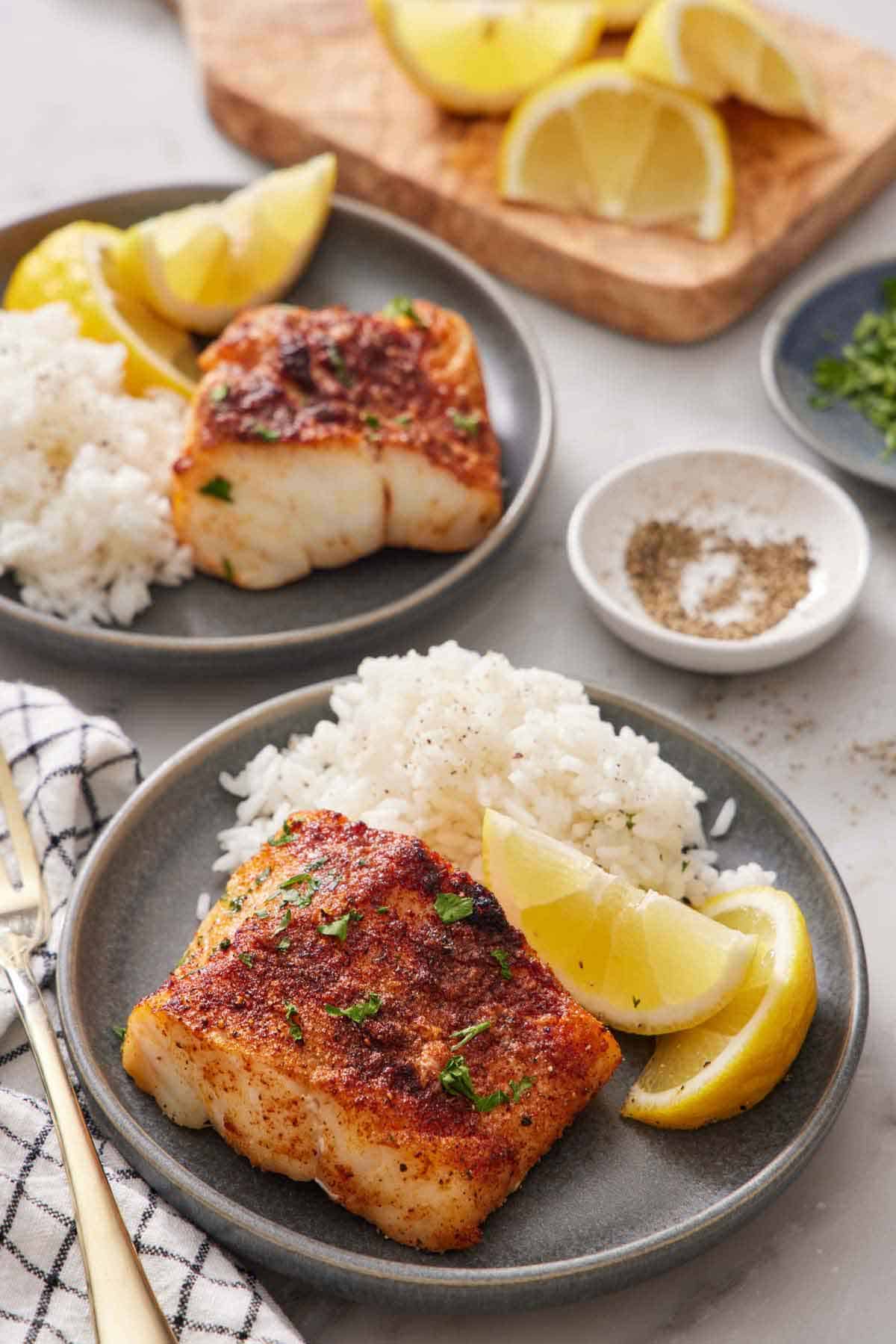 A plate with an air fryer cod, lemon wedges, and rice. A second plated portion in the back along with some pepper, parsley, and lemon wedges.