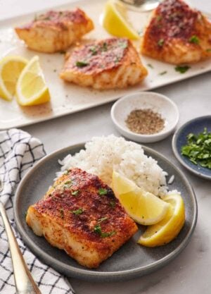 A plate with rice, lemon wedges, and an air fryer cod. A platter with more air fryer cod in the background along with lemon wedges. A bowl of pepper and parsley behind the plate.