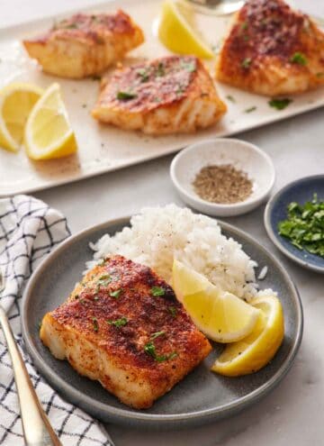 A plate with rice, lemon wedges, and an air fryer cod. A platter with more air fryer cod in the background along with lemon wedges. A bowl of pepper and parsley behind the plate.