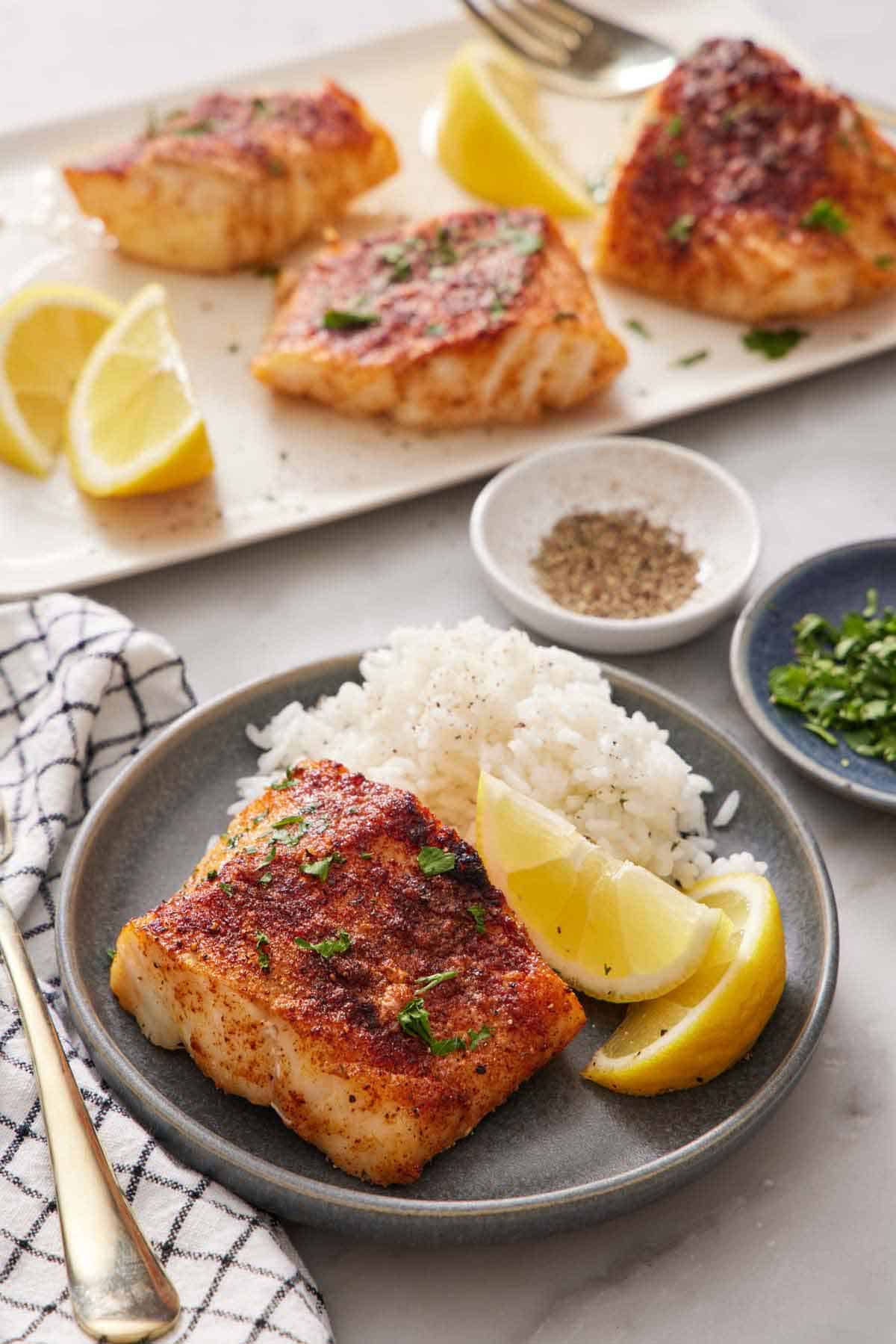 A plate with rice, lemon wedges, and an air fryer cod. A platter with more air fryer cod in the background along with lemon wedges. A bowl of pepper and parsley behind the plate.