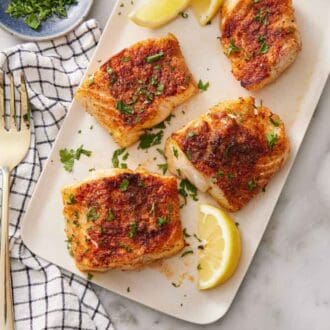 A platter of air fryer cod garnished with chopped parsley with lemon wedges. A fork and linen napkin on the side.