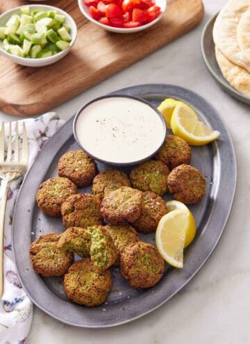 A platter of air fryer falafel with a bowl of dipping sauce and some lemon wedges. Pita bread in the background along with some cut tomatoes and cucumbers.