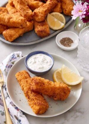A plate with two air fryer fish sticks with lemon wedges and a bowl of tartar sauce. A platter of more air fryer fish sticks in the background along with a bowl of pepper and a drink.
