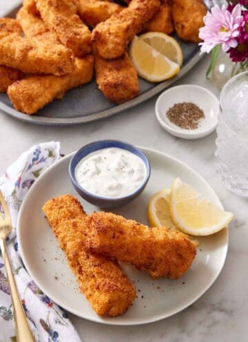 A plate with two air fryer fish sticks with lemon wedges and a bowl of tartar sauce. A platter of more air fryer fish sticks in the background along with a bowl of pepper and a drink.