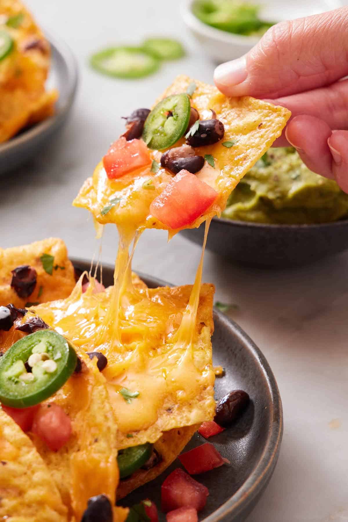 A tortilla chip with cheese, tomatoes, black beans, and jalapeno lifted from a plate of air fryer nachos.