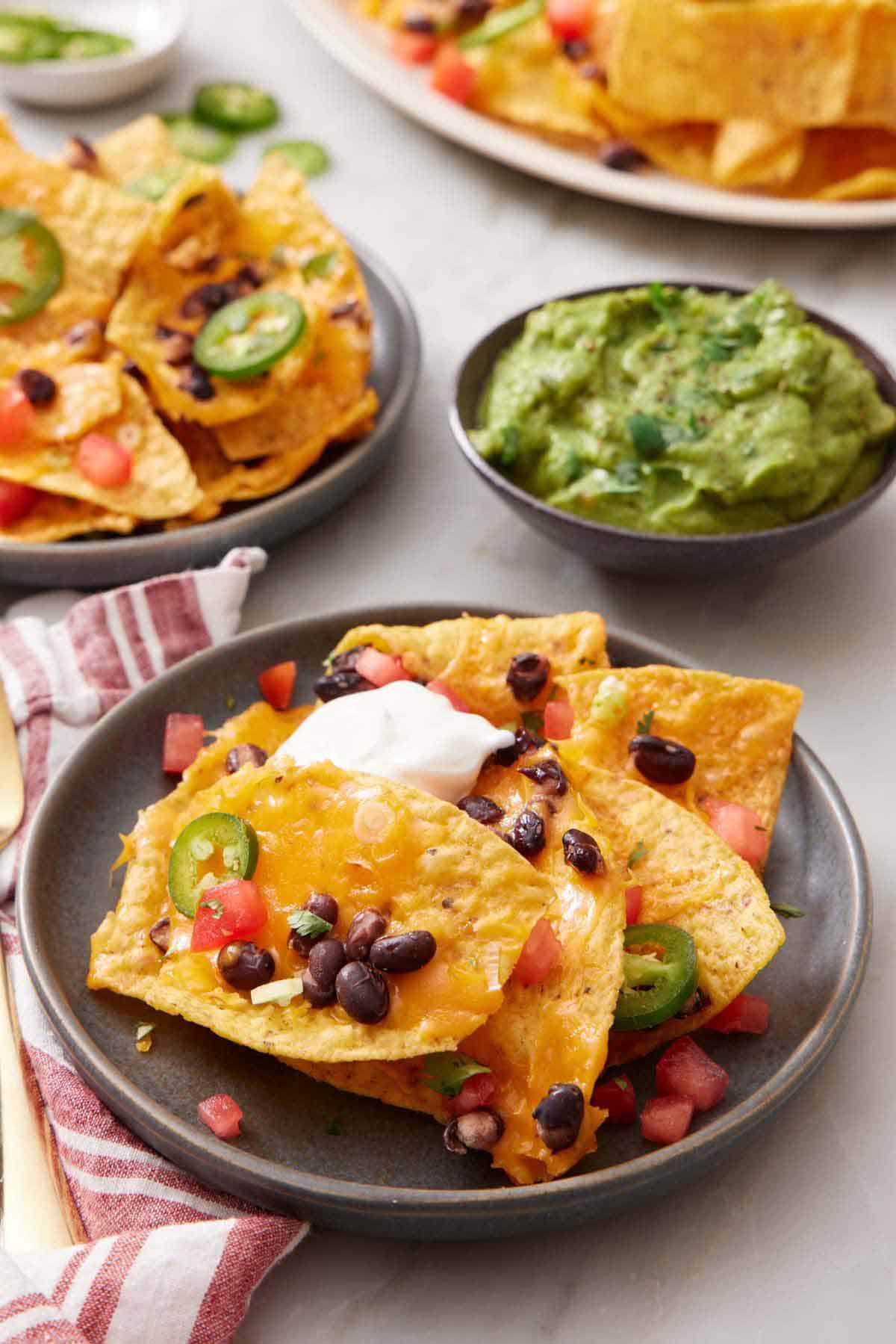 A plate of air fryer nachos topped with sour cream. A bowl of guacamole in the back along with more nachos.