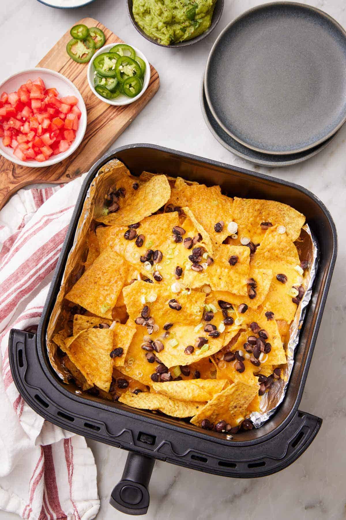 Overhead view of an air fryer basket lined with foil with air fryer nachos topped with green onions and black beans. A bowl of tomatoes, jalapeno, guacamole, and a stack of plates on the side.