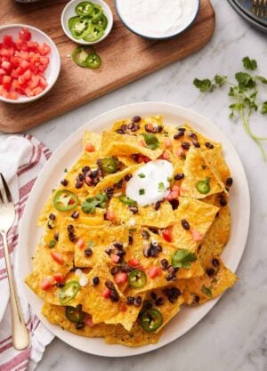 An overhead view of a platter of air fryer nachos with chopped tomatoes, jalapenos, cilantro, black beans, and sour cream. More toppings in bowls on the side.