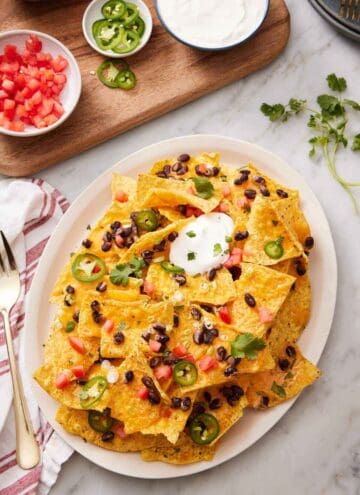 An overhead view of a platter of air fryer nachos with chopped tomatoes, jalapenos, cilantro, black beans, and sour cream. More toppings in bowls on the side.