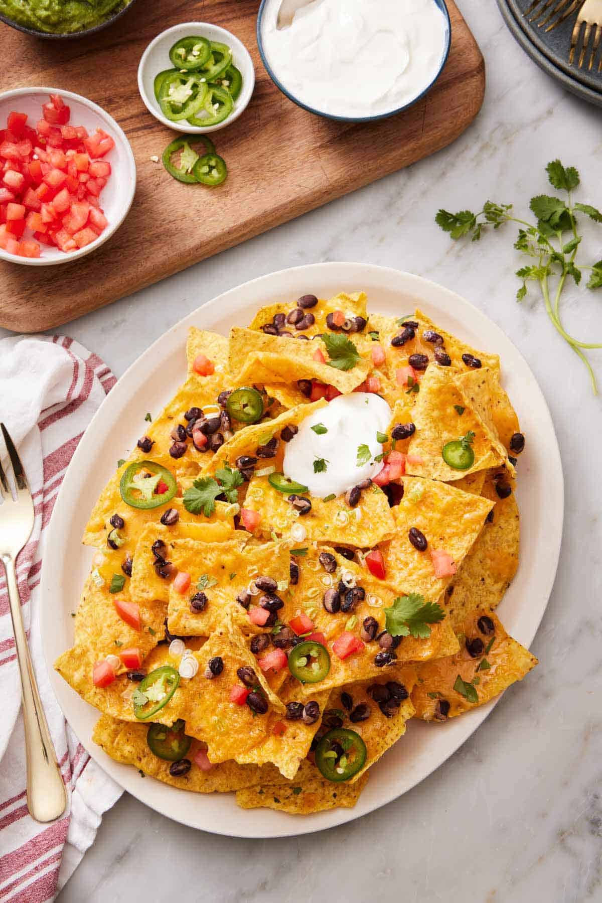 An overhead view of a platter of air fryer nachos with chopped tomatoes, jalapenos, cilantro, black beans, and sour cream. More toppings in bowls on the side.