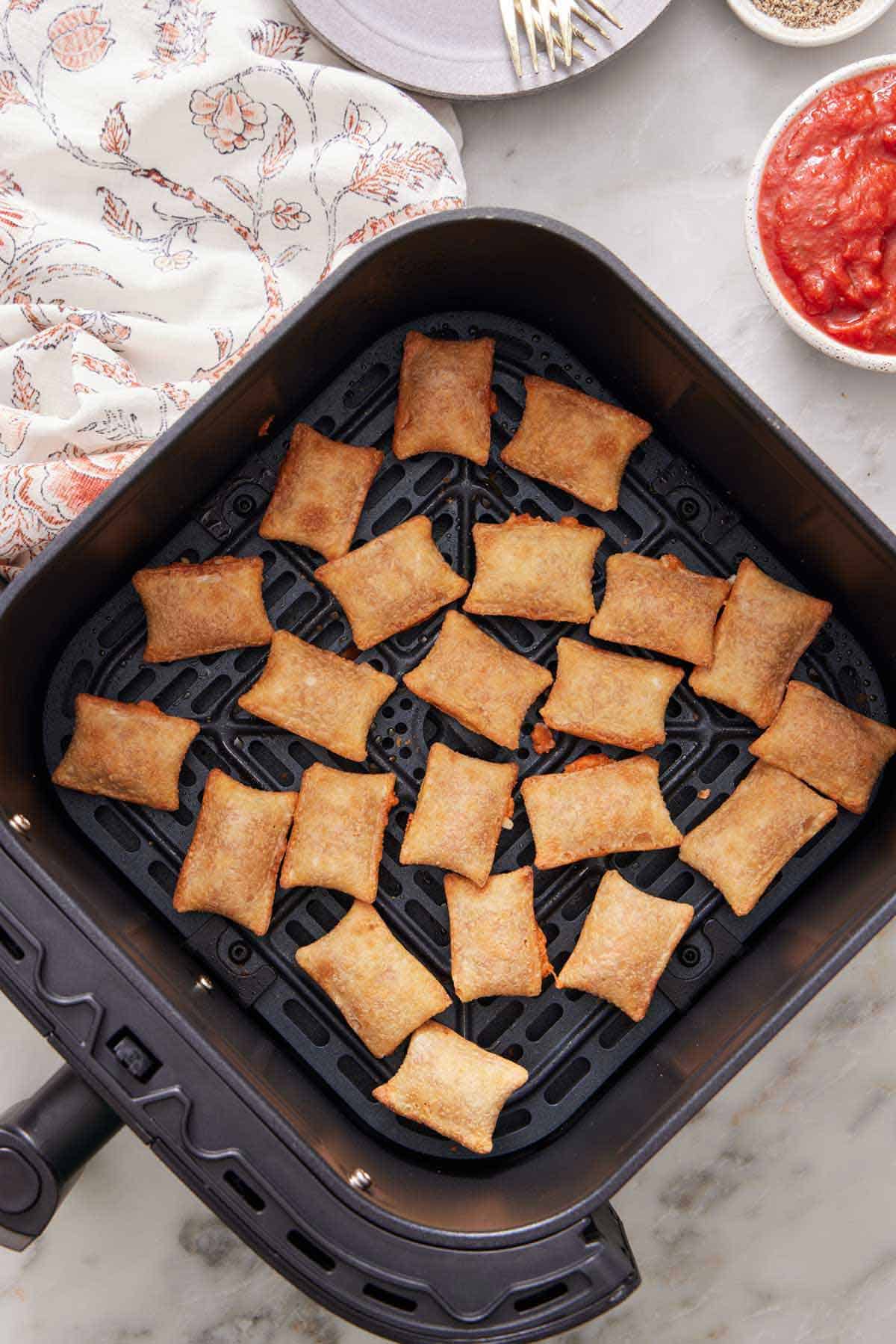 Overhead view of an air fryer basket with pizza rolls. A bowl of marinara on the side along with a linen napkin.