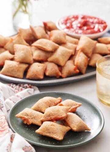 A plate of air fryer pizza rolls with a platter in the background containing more along with a bowl of marinara sauce.