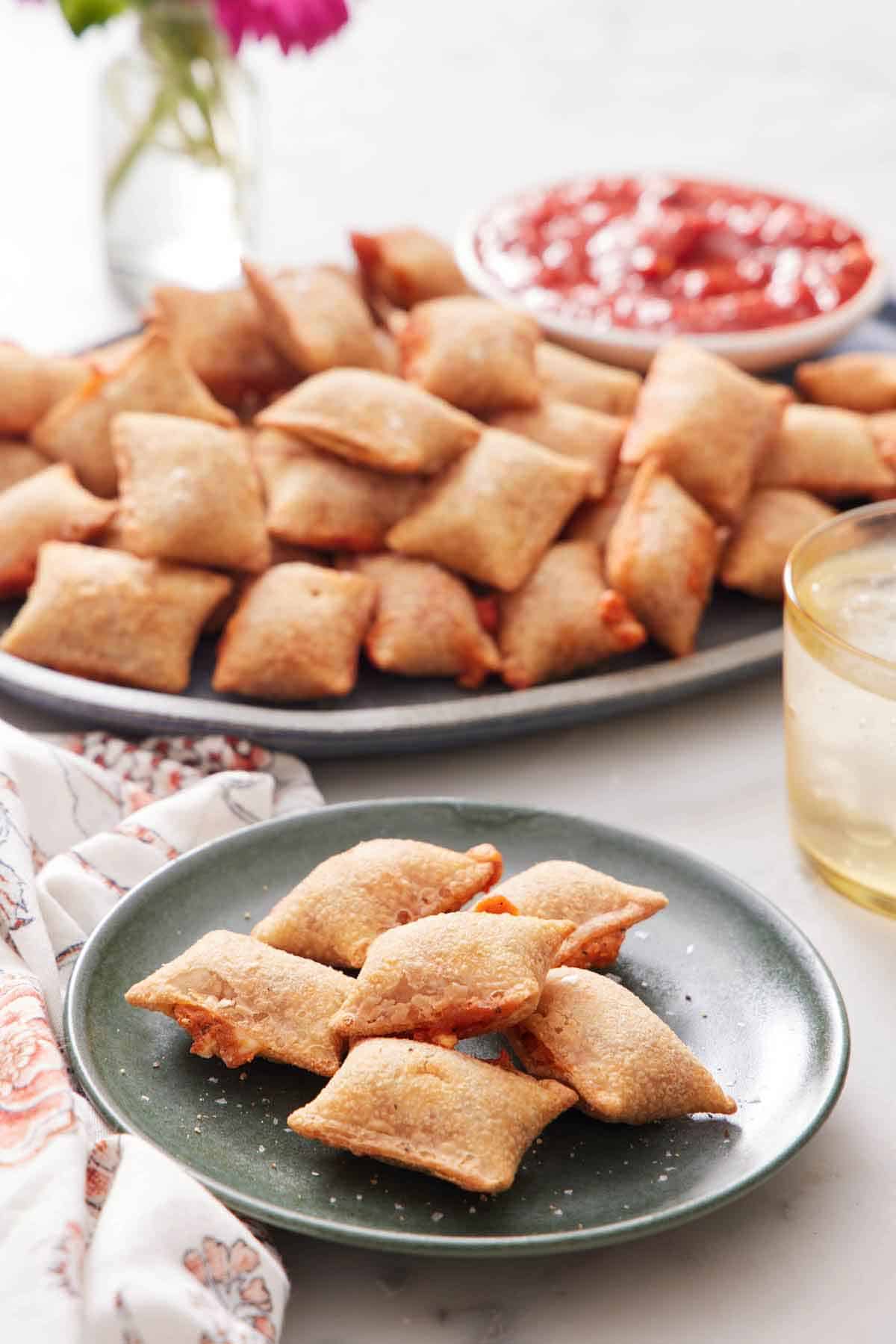 A plate of air fryer pizza rolls with a platter in the background containing more along with a bowl of marinara sauce.