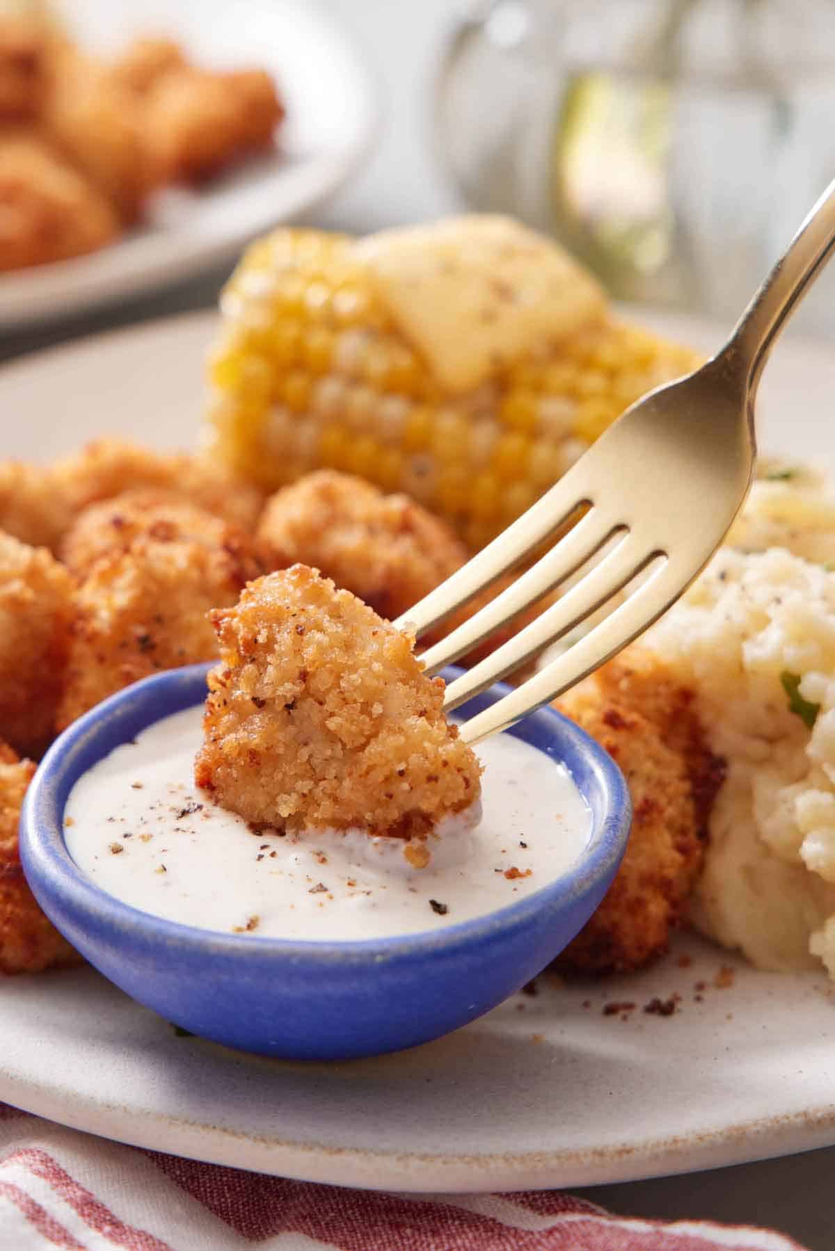 A fork dipping an air fryer popcorn chicken into a small bowl of sauce. Mashed potatoes, corn on the cob with butter, and more air fryer popcorn chicken on the plate.