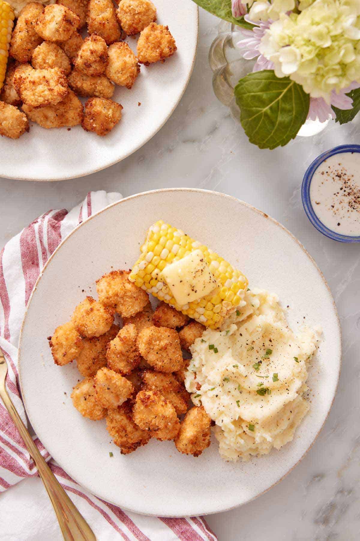 A plate with air fryer popcorn chicken, mashed potatoes, and corn on the cob with butter on top. More air fryer popcorn chicken on the side on a plate along with sauce and flowers.