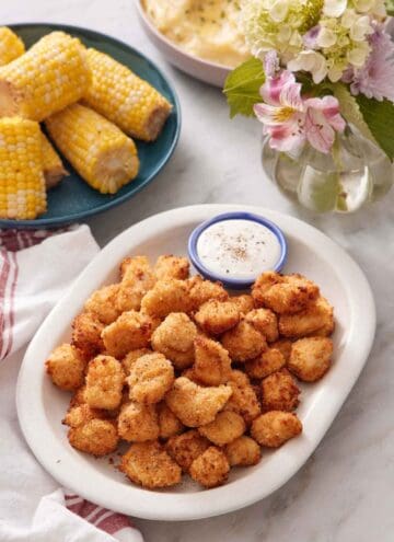 A platter of air fryer popcorn chicken with a bowl of sauce. Corn on the cob in the background along with flowers.