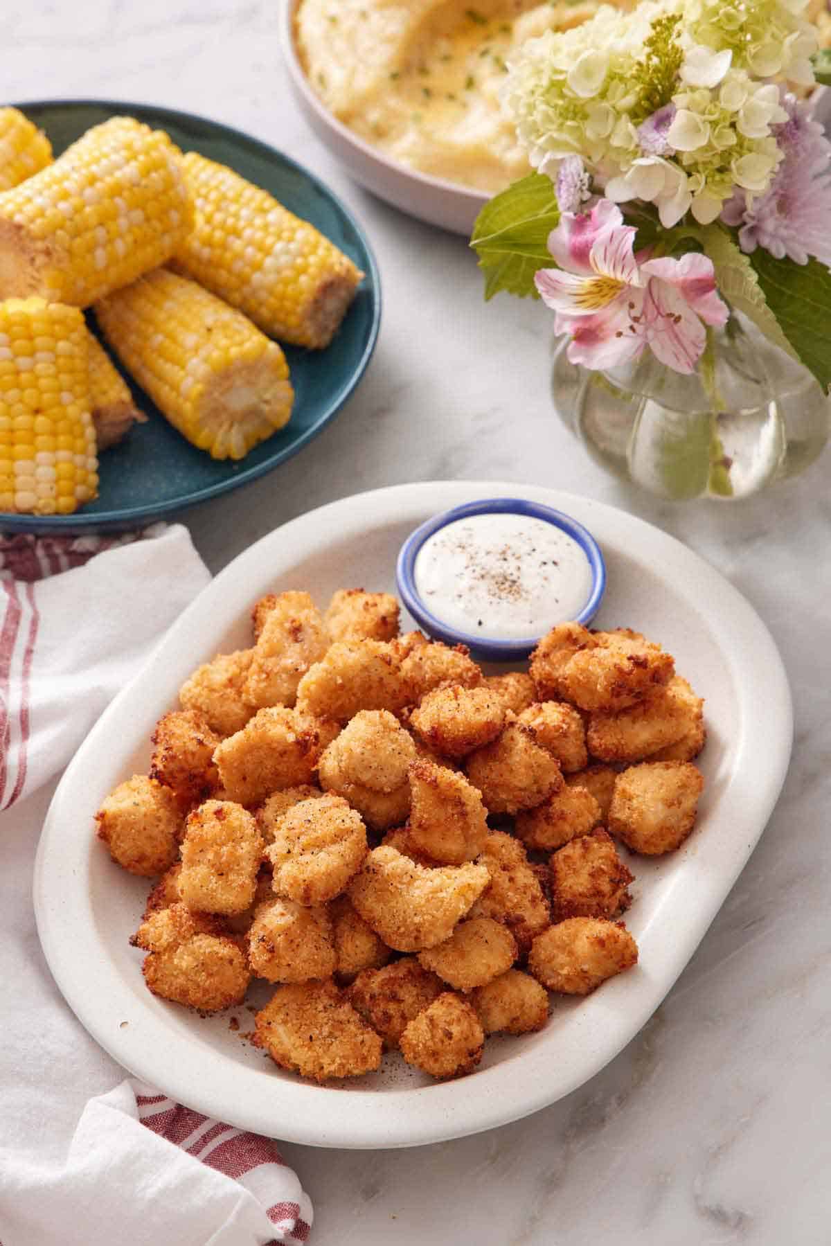 A platter of air fryer popcorn chicken with a bowl of sauce. Corn on the cob in the background along with flowers.