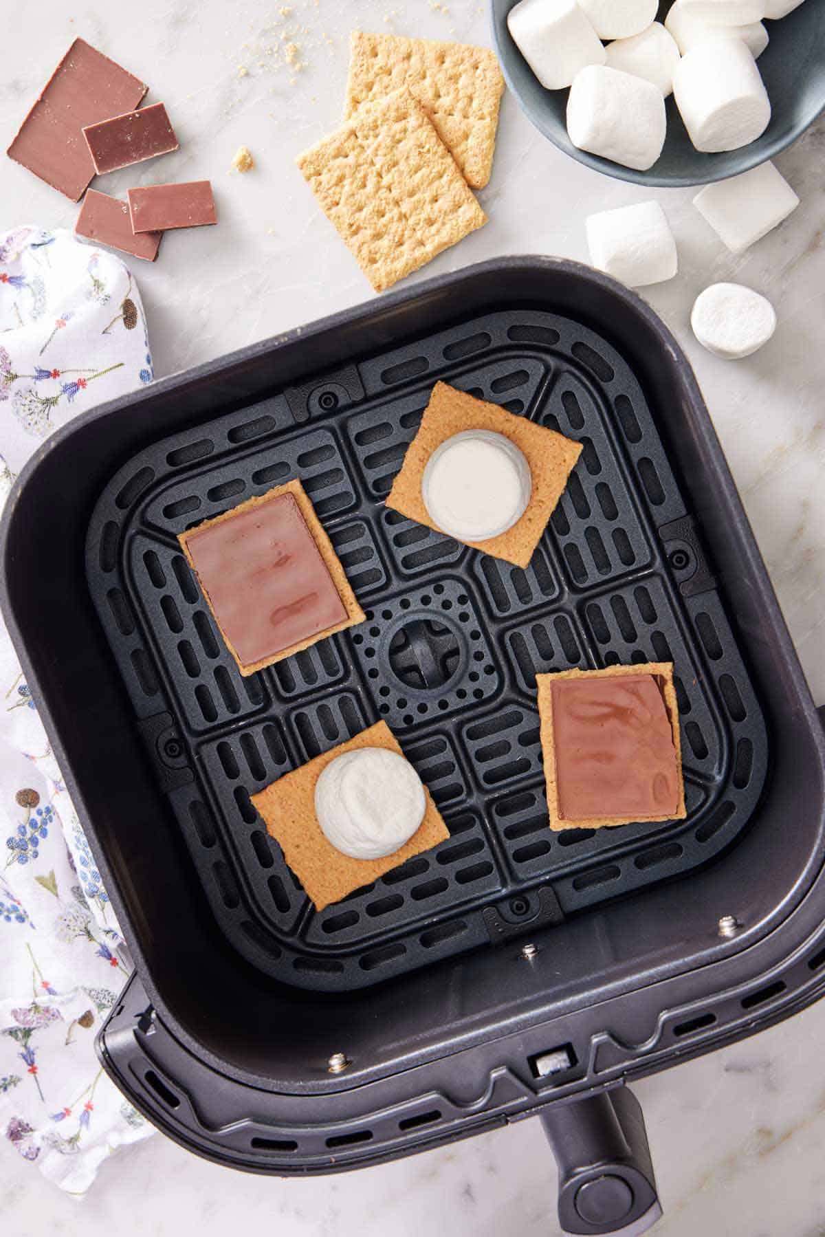 Overhead view of air fryer s'mores in an air fryer basket before air frying. Ingredients scattered around the back.