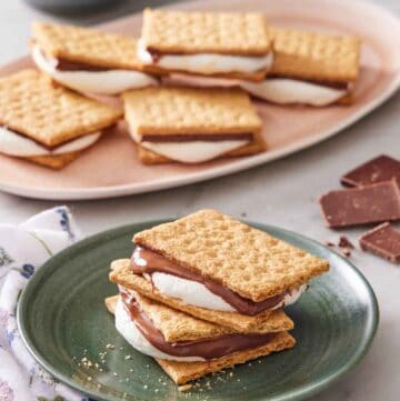 A plate with two air fryer s'mores stacked. More air fryer s'mores in the background on a platter with scattered chocolates and marshmallows.