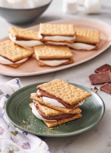 A plate with two air fryer s'mores stacked. More air fryer s'mores in the background on a platter with scattered chocolates and marshmallows.