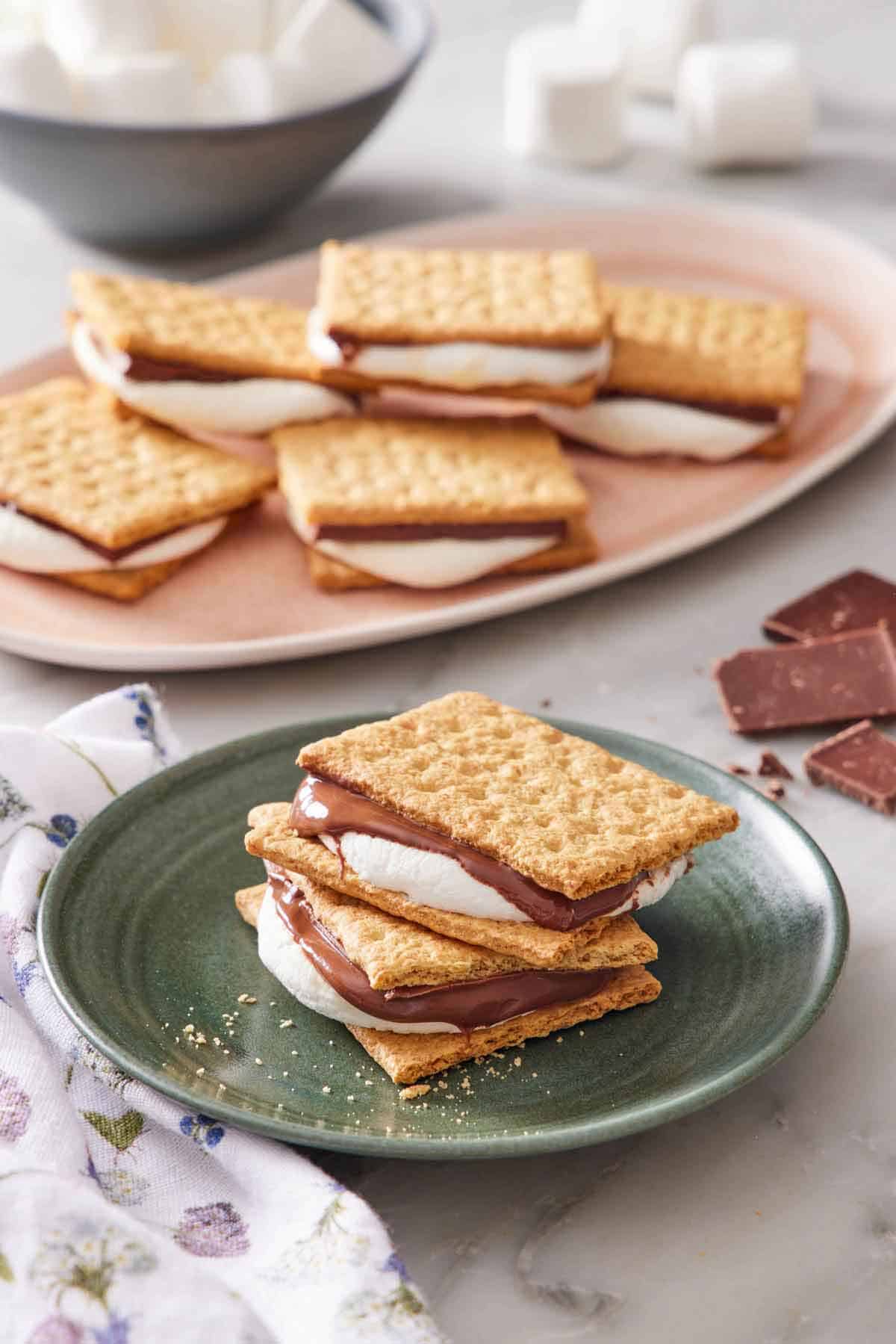 A plate with two air fryer s'mores stacked. More air fryer s'mores in the background on a platter with scattered chocolates and marshmallows.