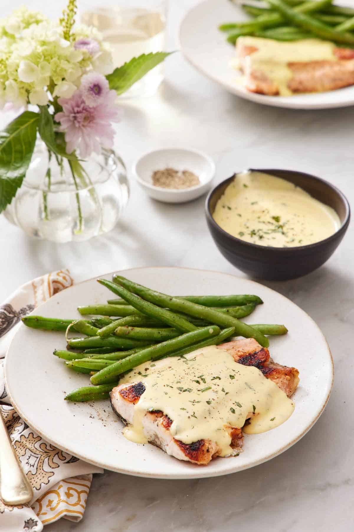 A plate of salmon with béarnaise sauce on top along with green beans. A bowl of béarnaise sauce in the background along with pepper and a vase of flowers.