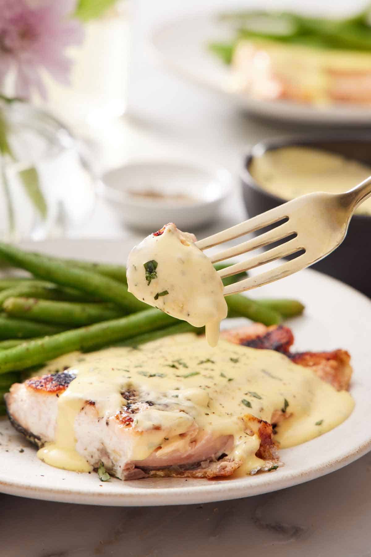 A fork lifting up a bite of salmon coated with béarnaise sauce from a plate with salmon and green beans.