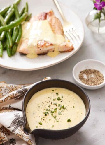 A bowl of béarnaise sauce with chopped tarragon leaves as garnish. A bowl of pepper in the background along with a plate of salmon and green beans with béarnaise sauce.