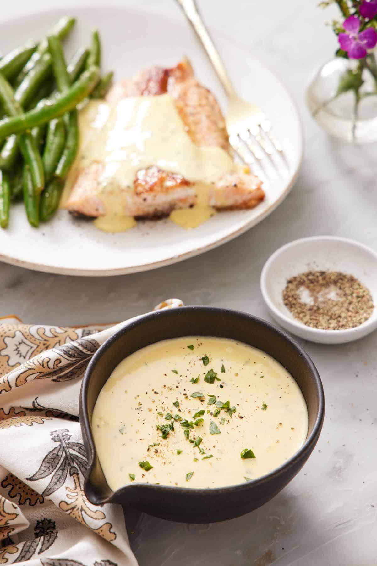 A bowl of béarnaise sauce with chopped tarragon leaves as garnish. A bowl of pepper in the background along with a plate of salmon and green beans with béarnaise sauce.