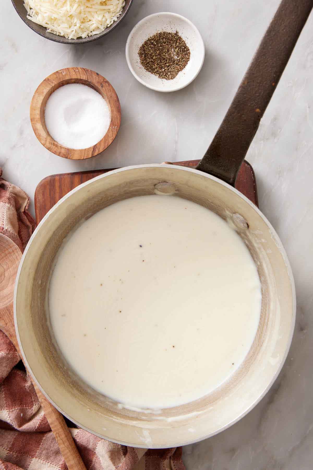 Overhead view of a pot of béchamel sauce. Salt, pepper, and shredded cheese in the background.