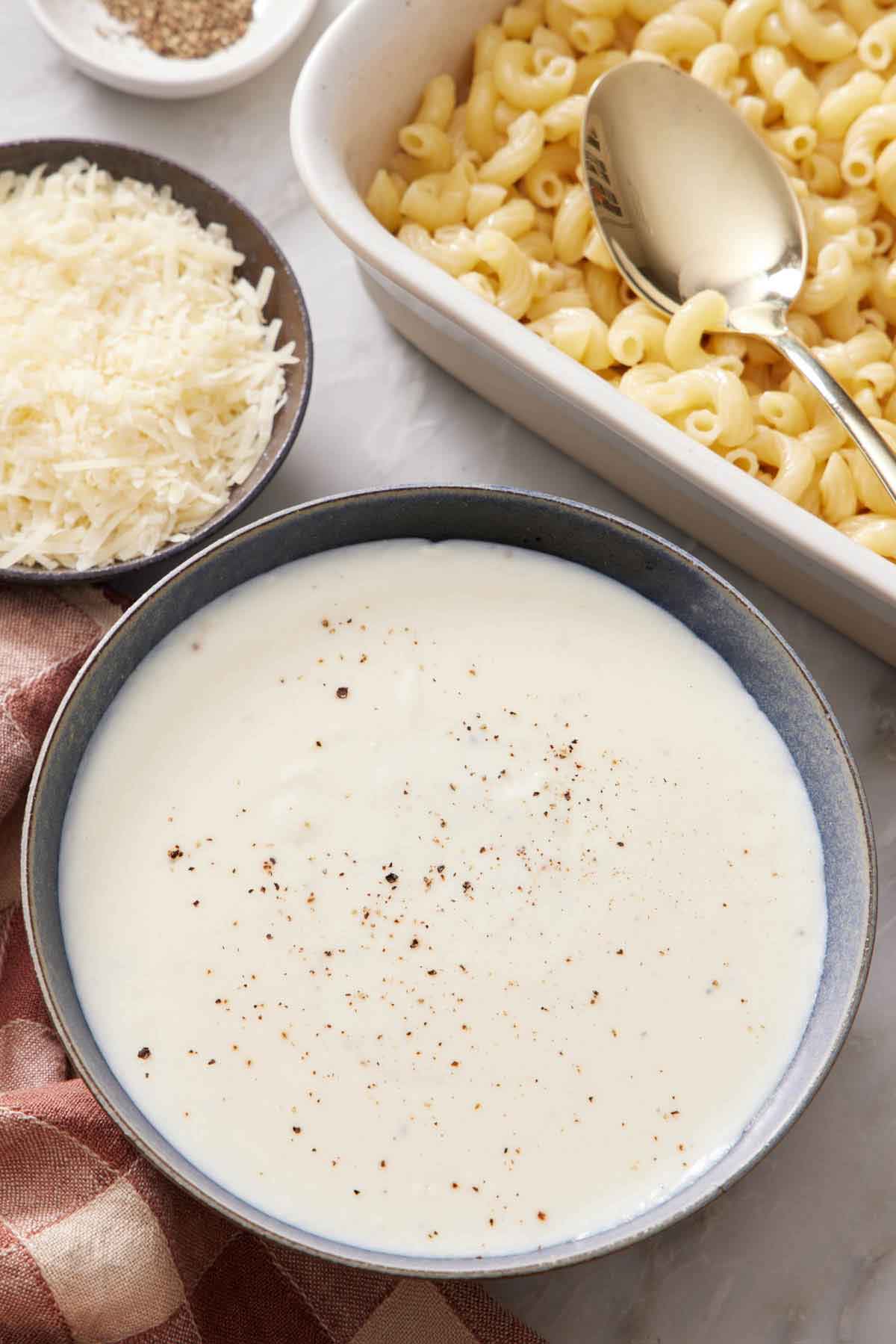 A bowl of béchamel sauce with pepper on top. Macaroni with a spoon in the back along with a bowl of shredded cheese.