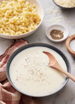 A bowl of béchamel sauce with a wooden spoon inside. Salt, Pepper, Cheese, and a bowl of macaroni in the background.