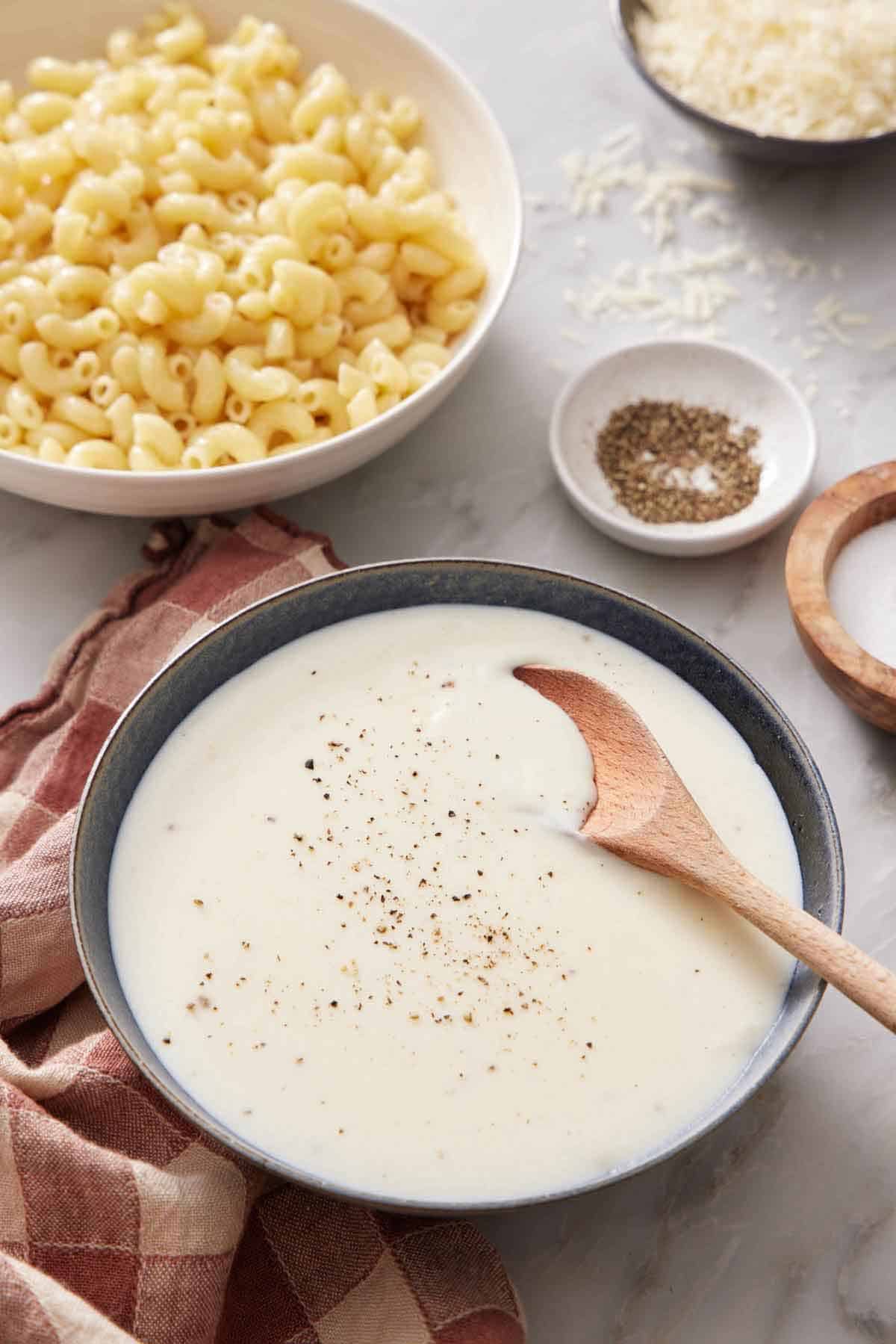 A bowl of béchamel sauce with a wooden spoon inside. Salt, Pepper, Cheese, and a bowl of macaroni in the background.