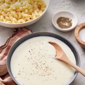 Pinterest graphic of a bowl of béchamel sauce with a wooden spoon inside. Salt, Pepper, Cheese, and a bowl of macaroni in the background.