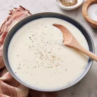 A bowl of béchamel sauce with pepper on top and a spoon inside. Salt and pepper in the background.