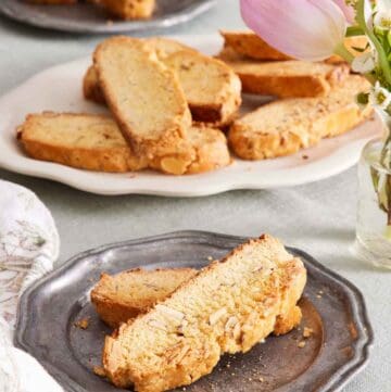 A plate with two biscotti. A platter in the background of more biscotti along with a plate with two more. Flowers to the side.