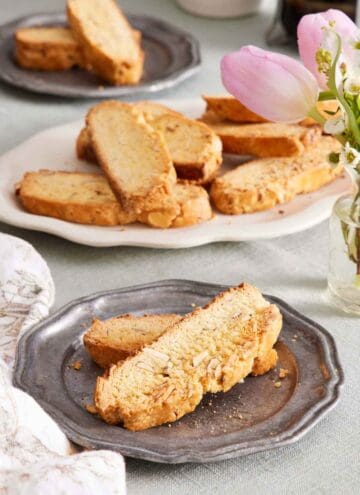 A plate with two biscotti. A platter in the background of more biscotti along with a plate with two more. Flowers to the side.