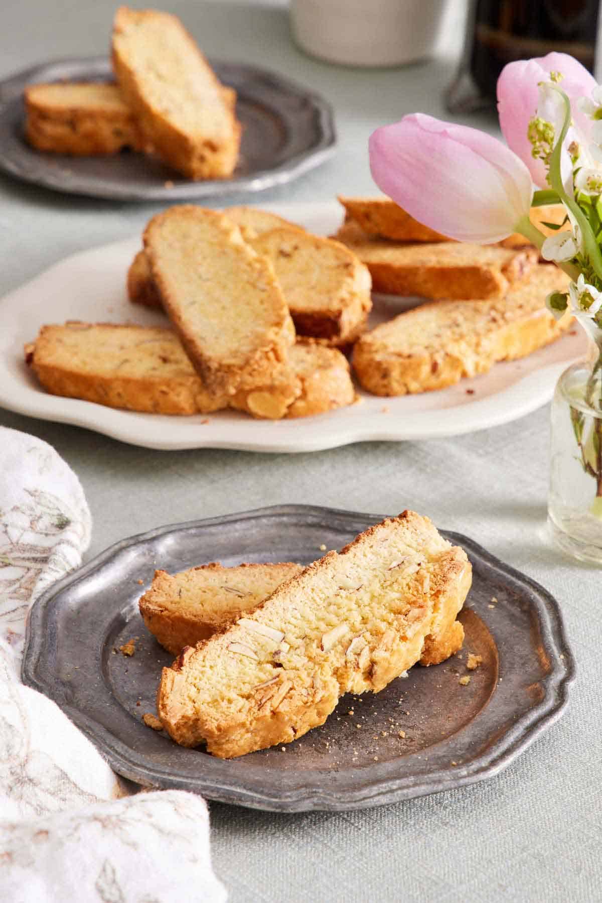 A plate with two biscotti. A platter in the background of more biscotti along with a plate with two more. Flowers to the side.