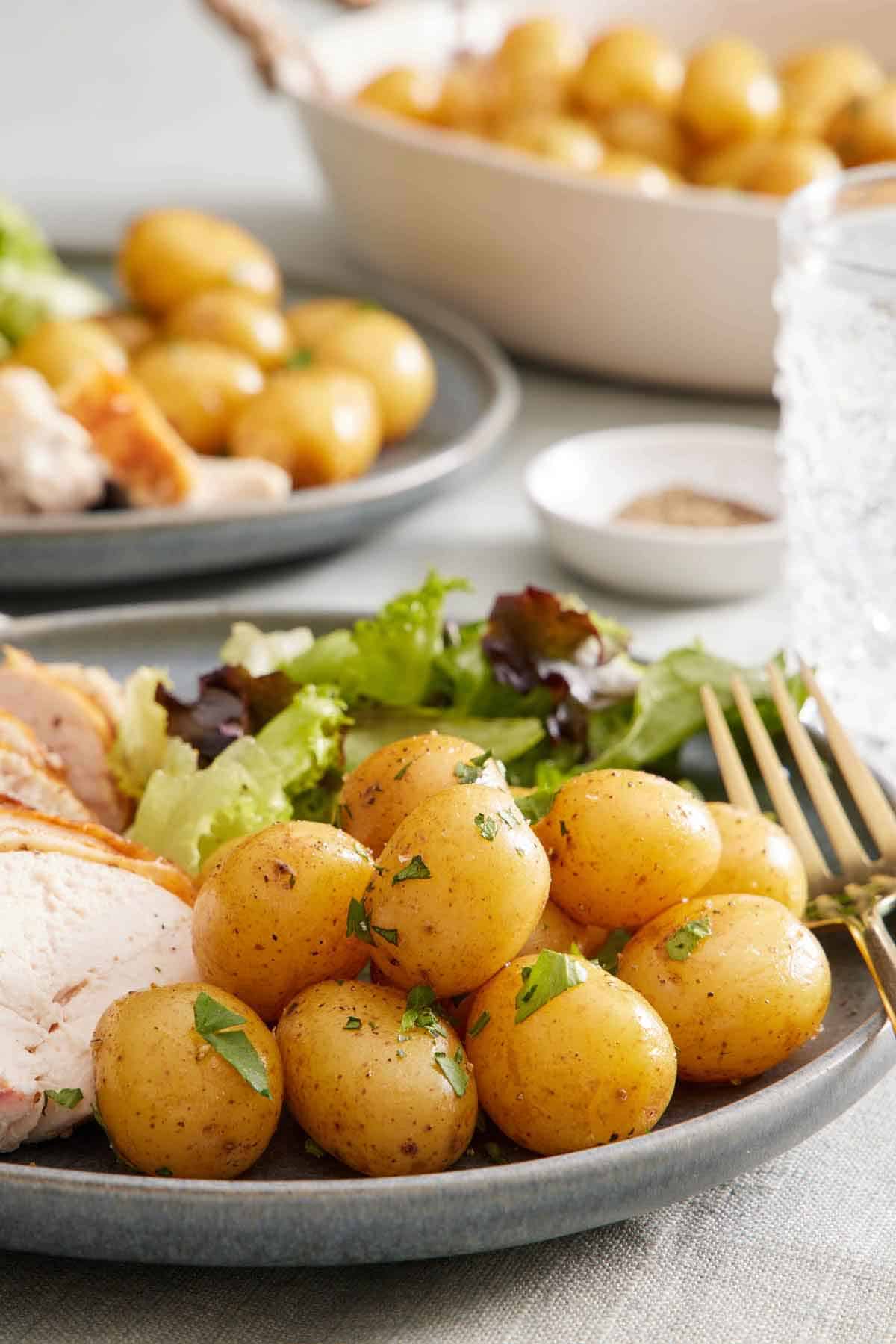 A close up view of boiled potatoes garnished with parsley on a plate with some mixed greens and chicken. Potatoes and another plated serving in the background.