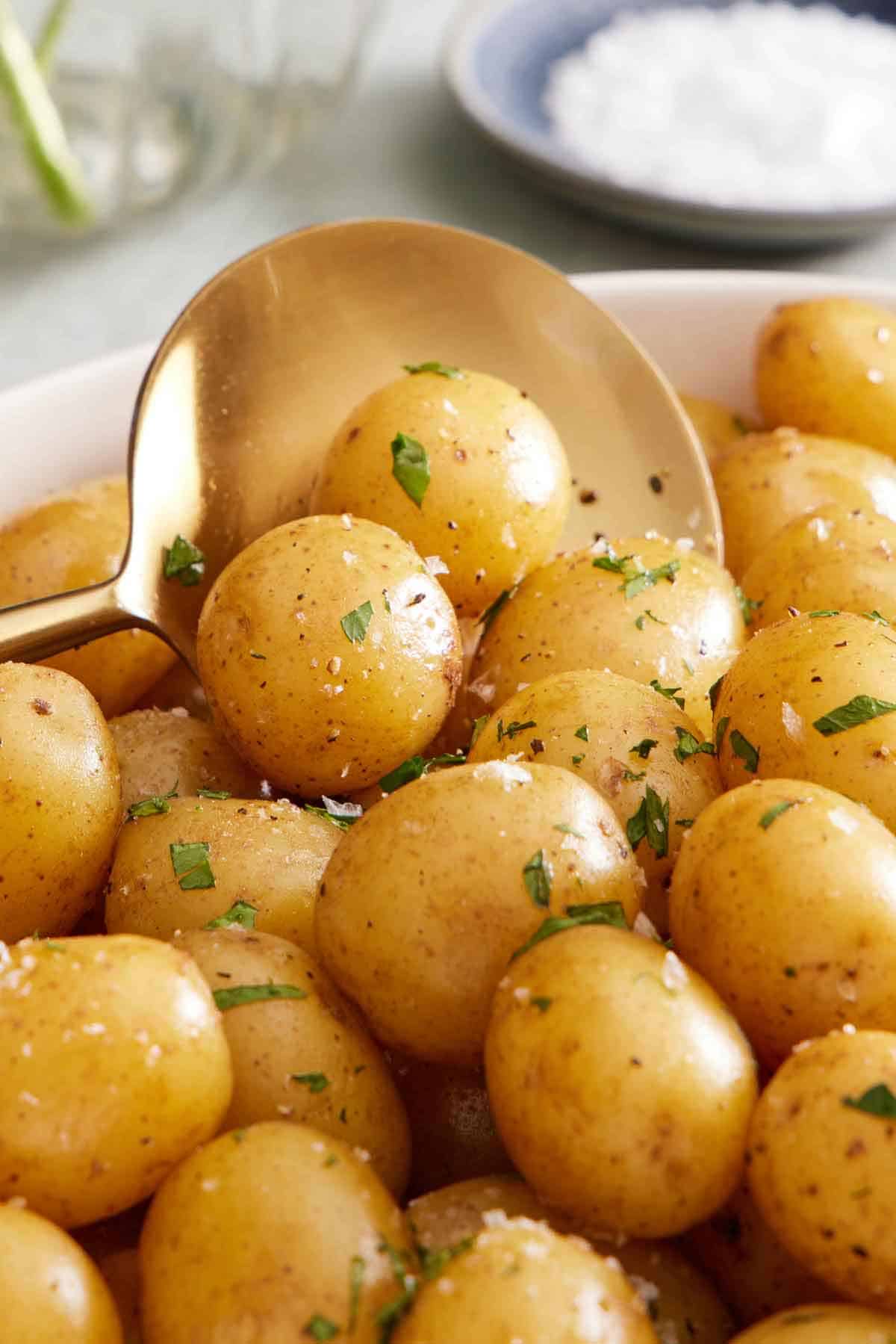 A close up view of a large serving soon scooping boiled potatoes with parsley, salt, and pepper.