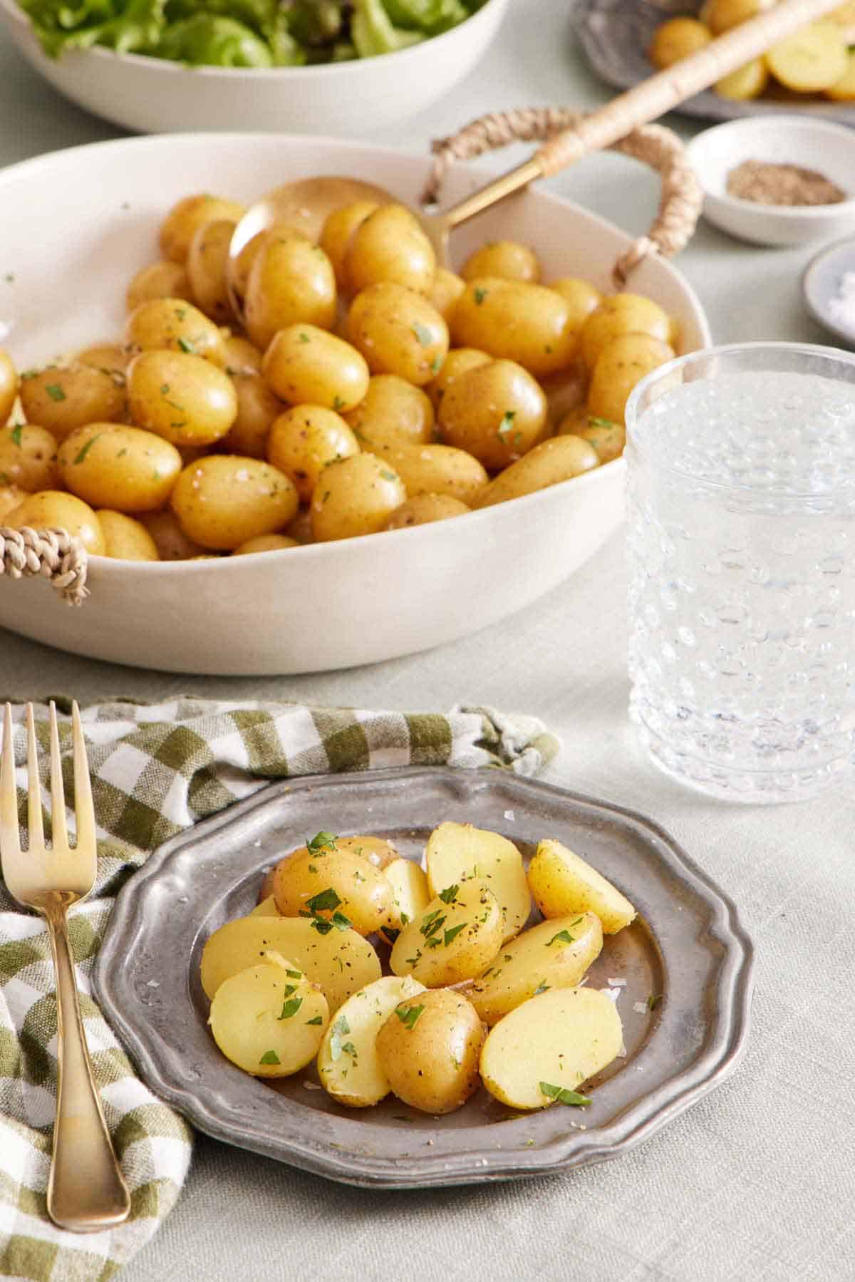 A plate with cut boiled potatoes. A drink in the background along with a large bowl of boiled potatoes.