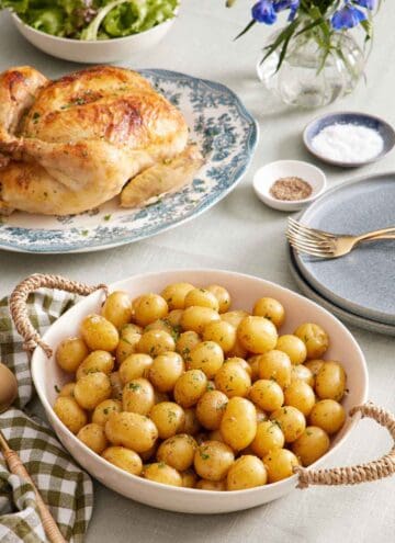 A large bowl of boiled potatoes. A roast chicken in the background along with plates, forks, salt and pepper, flowers, and some mixed greens in a bowl.