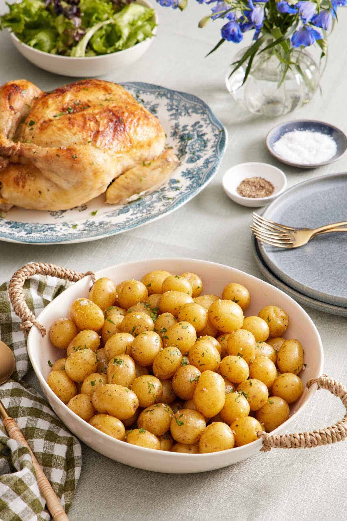 A large bowl of boiled potatoes. A roast chicken in the background along with plates, forks, salt and pepper, flowers, and some mixed greens in a bowl.