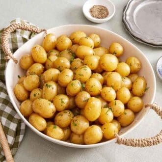 A large bowl of boiled potatoes garnished with chopped parsley. A napkin, serving spoon, salt, pepper, and plates on the side.