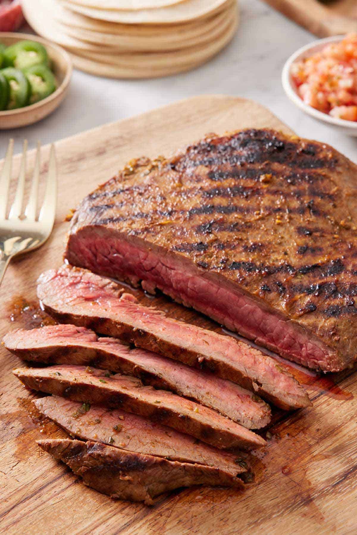 Carne asada on a wooden cutting board, partially sliced, showing the interior. A fork and taco toppings in the background.