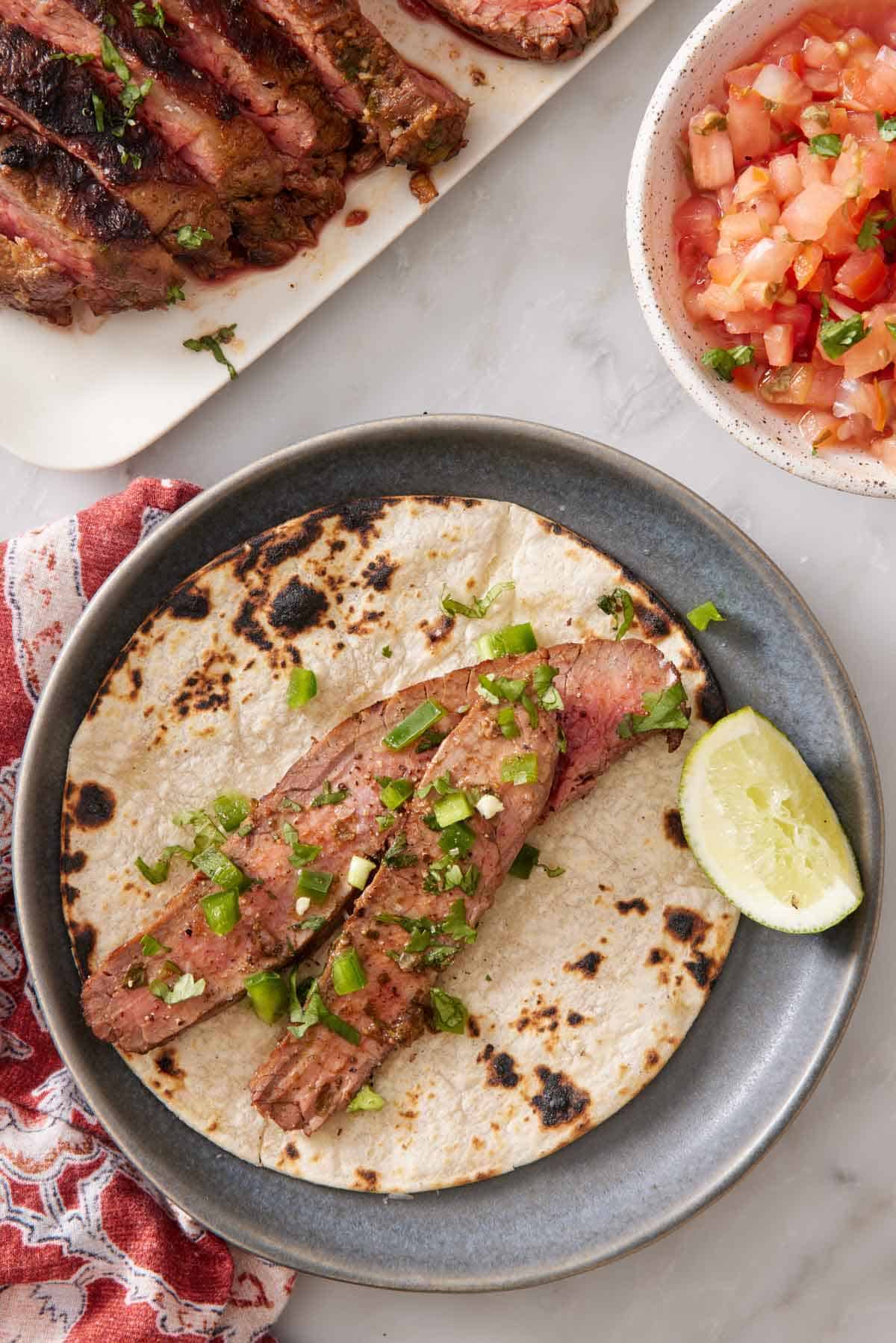 Overhead view of a corn tortilla with sliced carne asada topped with chopped cilantro and a lime wedge. Pico de gallo and more carne asada off to the side.