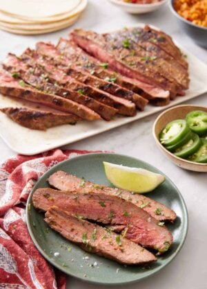A plate of carne asada with a lime wedge. A platter with the rest of the carne asada in the background also with tortillas, sliced jalapenos, and rice.