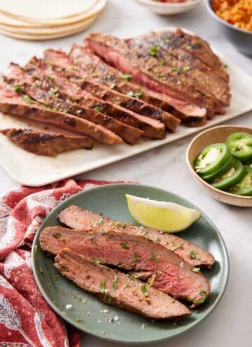 A plate of carne asada with a lime wedge. A platter with the rest of the carne asada in the background also with tortillas, sliced jalapenos, and rice.