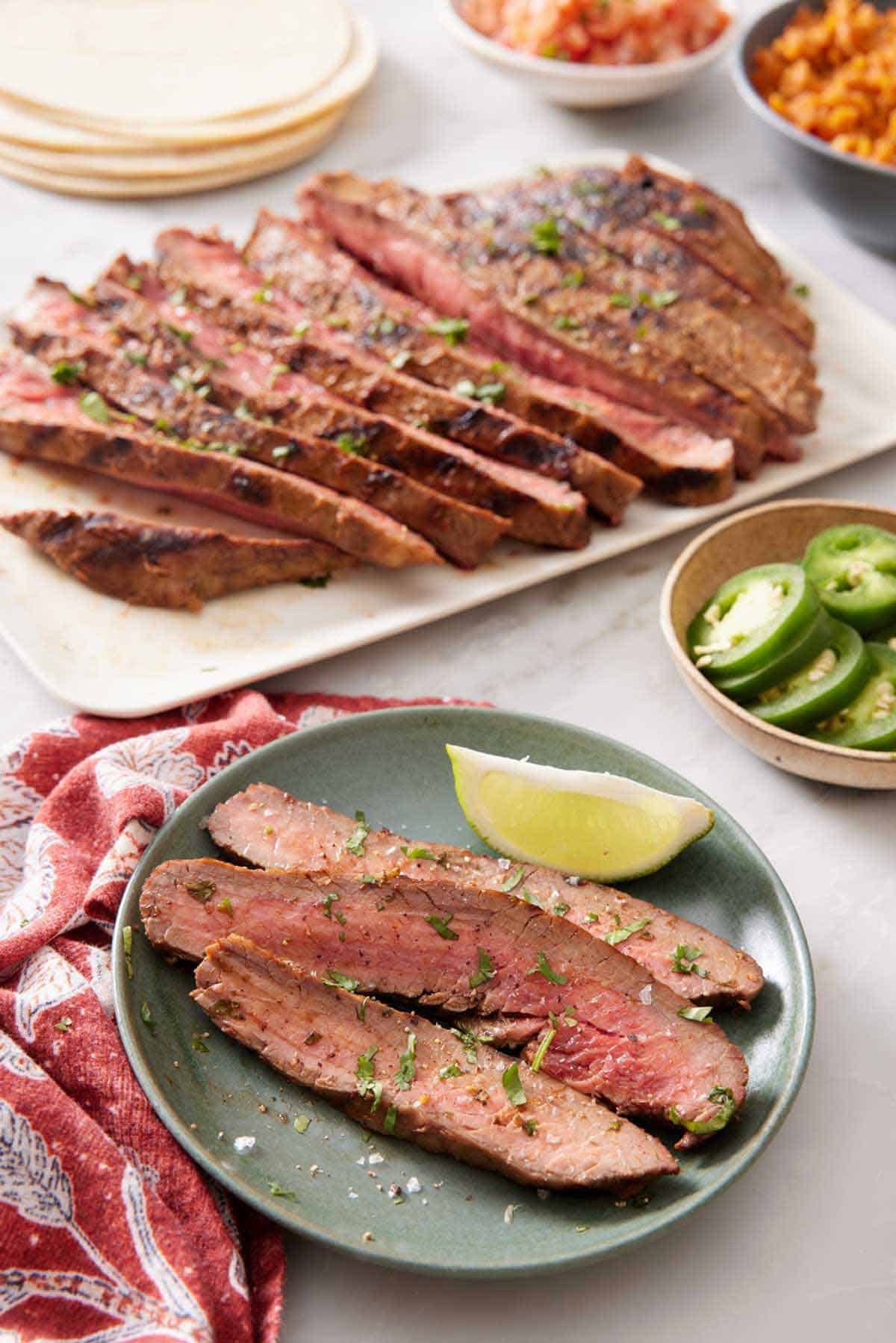 A plate of carne asada with a lime wedge. A platter with the rest of the carne asada in the background also with tortillas, sliced jalapenos, and rice.