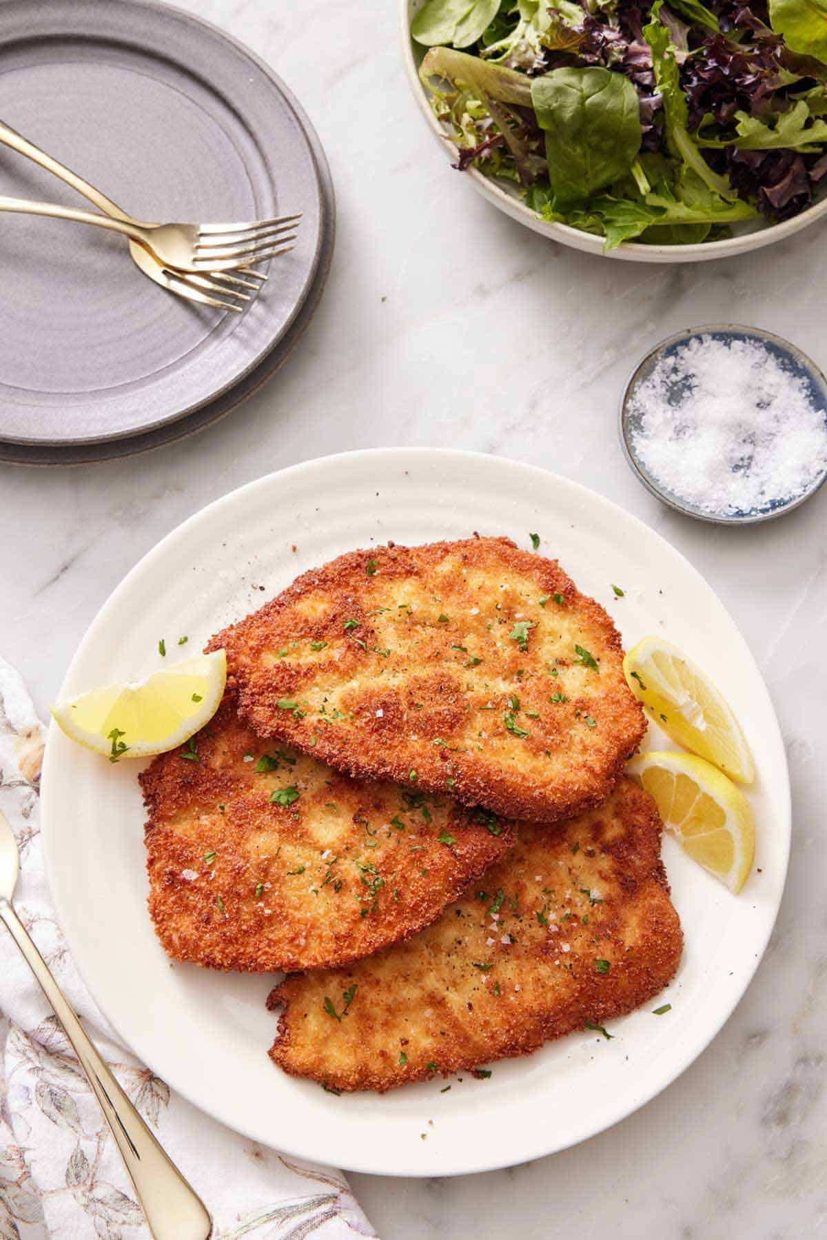 Overhead view of a platter of chicken schnitzel with lemon wedges. A small bowl of salt, a stack of plates and forks, and a bowl of mixed greens on the side.